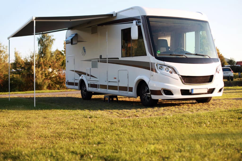 Camping-car intégral blanc garé sur l'herbe, avec son grand auvent gris déployé. Lumière du soleil couchant.