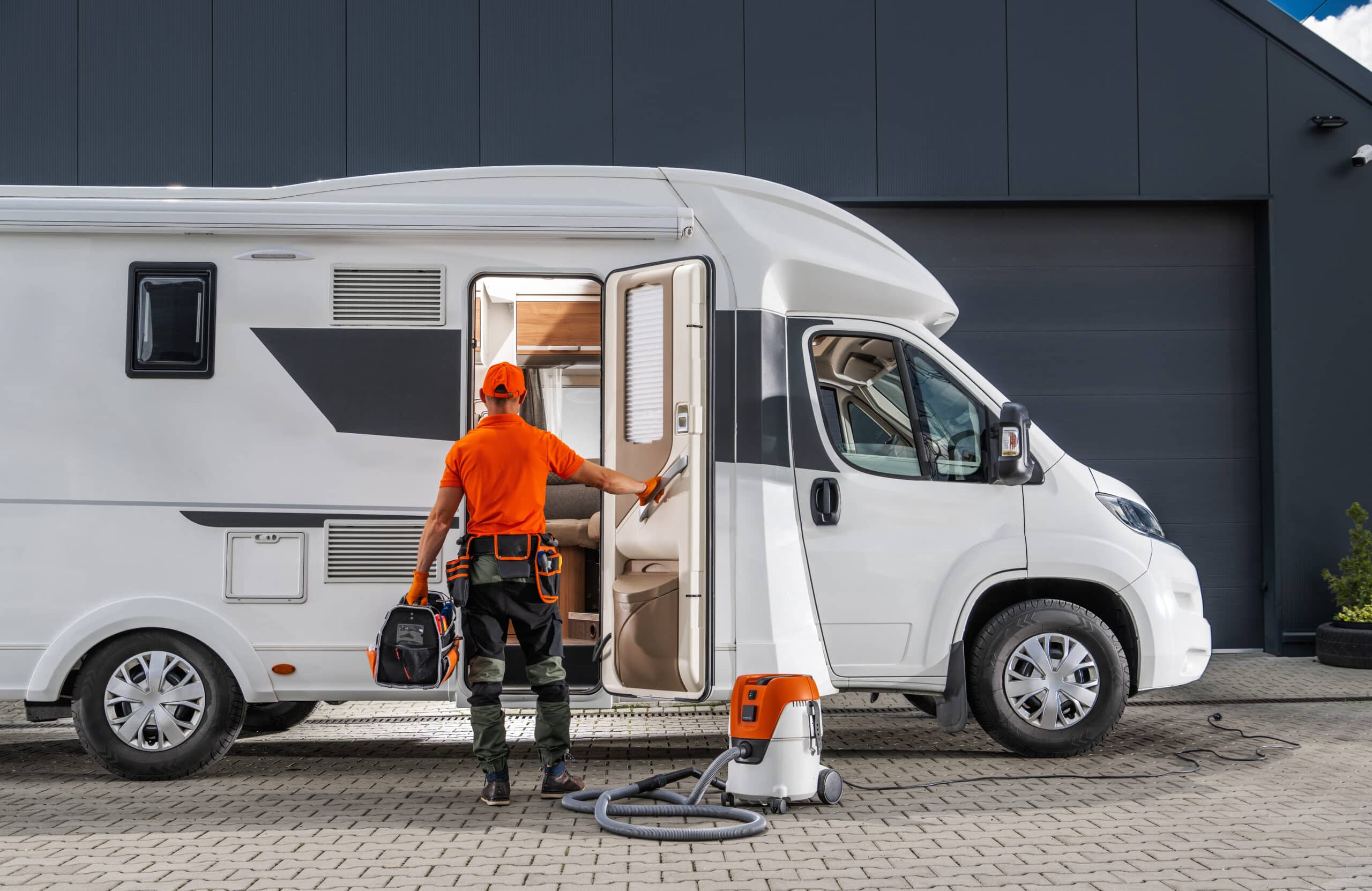 Technicien en orange prêt à l'entretien d'un camping-car blanc. Il tient sa trousse d'outils; un aspirateur professionnel est au sol.