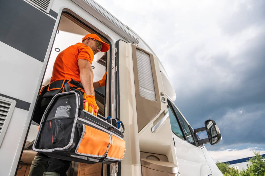 Technicien VR: Maintenance et réparation de camping-car. Technicien VR en orange vif entre dans un camping-car blanc. Il porte des gants, lunettes de sécurité et un sac à outils.