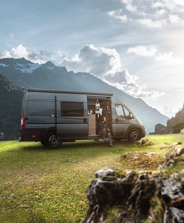 Van aménagé en montagne : aventure et confort en nature Camping-car aménagé gris garé dans une prairie face à de hautes montagnes enneigées, une femme se repose à l'entrée.